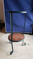 Front and angled view of a two-shelf plant holder with round wooden shelves and green metal frame with curly legs.