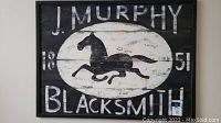 Close-up photo of the wooden black and white sign reading 'J Murphy Blacksmith 1851' featuring a galloping horse.