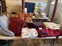 Table with various blankets, throws, and pillows including weighted blanket, red plaid throw, patterned pillows, cream comforter, and multicolored throws.