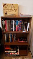 Photo of medium-brown wooden bookcase filled with assorted books on two lower shelves and magazines and calligraphy kit on top shelf
