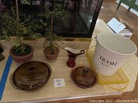 View of two brown tinted glass casserole dishes with lids, two artificial plants in brown pots, red ceramic milk/cream dispenser, white metal trash bucket with black 