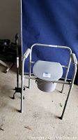 Folding metal frame commode with arms, seat, lid, and removable bucket below, with quad cane standing beside it.