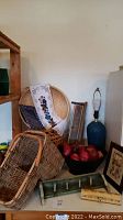 Wide angle photo showing assortment of baskets, metal bowl with apples, lamp base, small shelves and framed art pieces.