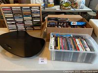 Photo showing wooden rack with cassette tapes, plastic tub with multiple DVDs, box with DVD TV show sets, and portable black lap desk on table.