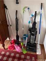 Various cleaning supplies grouped together including upright vacuums, dusters, spinners, and hardwood floor cleaning tools.