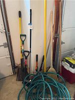 Photo showing shovels, rake, broom, green garden hose, and Fiskars hedge shears grouped standing upright against a wall in garage.
