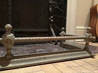 Front view of brass fireplace fender showing twisted rope-style bar and decorative end posts.