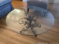 Top-down view showing clear round glass top and ornate metal base on hardwood floor