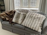 Three large animal print faux fur pillows and a leopard print throw displayed on a gray sofa in a living room setting with window and curtains in background.