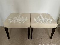 Pair of beige linen upholstered ottomans side by side showing chandelier print and dark wood legs.
