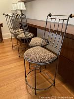 Four metal bar stools with pewter colored metal frames and leopard print velvet cushions shown at desk height. Burnished brass knob accents on backrests visible. Hardwood floor and cabinetry in background.