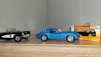 Blue hardtop Corvette model with chrome details, next to black convertible with police badge and clear windshield, both on wooden surface.