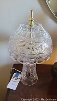 Full view of a clear cut glass lamp on a wooden surface with brass finial and detailed cut floral and geometric design on glass.