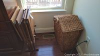 Photo of the woven rectangular storage basket placed by a wall near a window on hardwood floor. The basket is natural fiber colored light brown with visible wear.