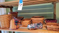 Shelf with various Longaberger baskets without handles in different sizes and shapes, some with fabric liners and one cone-shaped basket with plaid bow.