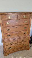 Front view of wooden chest of drawers with brass handles, showing multiple drawers and decorative top edge.