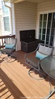 Photo showing two patio swivel chairs, part of round glass-top table, and the brown storage bin on a wooden deck.