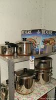 Shelf with an assortment of pots and pans including stainless steel stockpots, nested saucepans and copper-bottomed pots, with Revere Stainless Steel box in background.