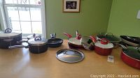 Overview of red and black pots and pans with lids on a light wood table in front of a window and green walls.