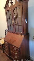 Full view of wooden secretary desk showing closed glass paneled upper cabinet, ornate top, and lower slant-front desk with drawers.