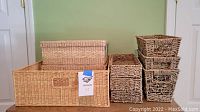 Nine woven storage baskets arranged on a wooden surface. Large rectangular basket at bottom with medium basket on top left and six smaller baskets stacked on right.