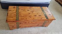 Closed wooden storage trunk with metal strap accents on top and sides, placed on carpeted floor.