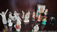 Front view showing 12 off-white Willow Tree figurines with wire wings and 6 colorful folk art figurines, plus a small framed picture on the right.