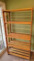 Photo of a wooden shelving unit with five shelves placed against a green wall near a glass door, showing the full height and construction of the unit.