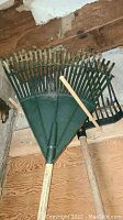 Multiple green plastic head garden rakes with wooden handles shown resting against wall