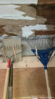 Photo showing a wooden rake, metal pitch fork, and a blue plastic leaf rake all with wooden handles, set against a wooden wall.
