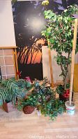 Artificial plants including large tree, smaller plants, ivy garland, and bamboo coat rack arranged on floor near wall with sunset silhouette.