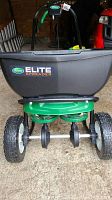 Front view showing Scotts Elite logo on the large black hopper and visible dual spinning discs below the hopper. Large pneumatic tires provide mobility.