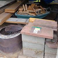 Overview of stacked cement blocks with cement bricks/pavers on top, green wheelbarrow behind with wood scraps, perforated burning barrel with grill grate in front