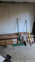 Photo showing wood trim pieces stacked along wall with green broom, shovels, and hand saws leaning against wall