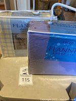 Two packaged twin flannel sheet sets, one lavender and one with plaid packaging, sealed with rope handles visible