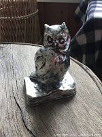 Front and side view of a chrome owl hood ornament sitting on a wooden surface with natural light highlighting the detailed features and texture.