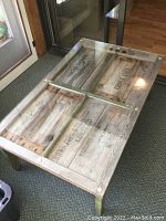 Rectangular coffee table made from antique wooden crates with a clear glass top, showing rustic wood panels and painted legs.