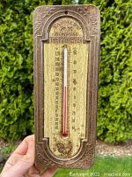 Front view of the antique tin thermometer held in hand outdoors, showing embossed details, temperature scales and red liquid column.