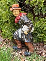 Side view of the folk art statue showing the face, orange hat, red scarf, black coat, and brown pants in outdoor greenery setting.