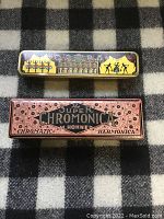 Photo of two harmonicas with closed cases, one Opera branded with colorful tin case, one Hohner Super Chromonica with printed box.
