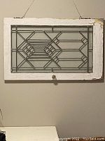 Front view of antique leaded glass window hung on wall showing geometric lead design and white wooden frame with chipping paint.