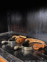 Pair of metal cowboy spurs with custom brown leather straps, displayed on a dark wood surface.