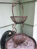 Front view of the antique rustic iron plant stand showing two open basket holders and curved top handle resting against a green wooden wall.