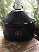 Front view of black painted metal traveling box with conical lid, oval handle, and painted insignia.