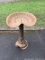 Full view of the antique tractor seat stool showing the metal seat, cylindrical post, and base on outdoor pavement with some grass in the background.