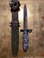 Antique military cutter knife displayed alongside original green metal sheath with brown woven strap and metal fasteners on a wood surface.