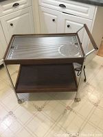 Full view of mid century modern hot plate trolley showing chrome handles and frame, electric hot plate top with geometric design, teak wood bottom shelf, and caster wheels.