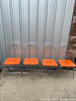 Front view of four folding chairs with chrome frames and orange vinyl seats, placed side by side.