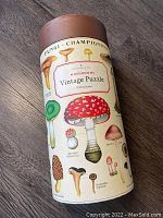 Cylindrical box of 1000 piece vintage mushrooms jigsaw puzzle showing botanical illustrations of various mushrooms on cream colored background with brown lid.