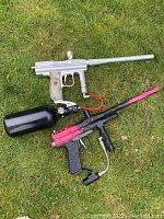 Two vintage paintball guns laid on grass, one silver with black grip and another black and red with air tank.
